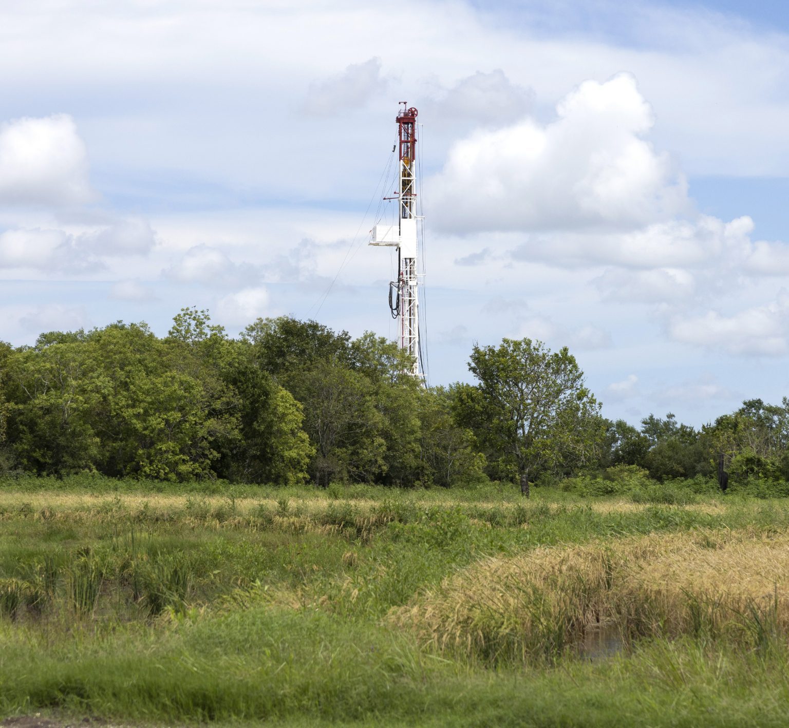 An oil rig behind a green landscape and trees.